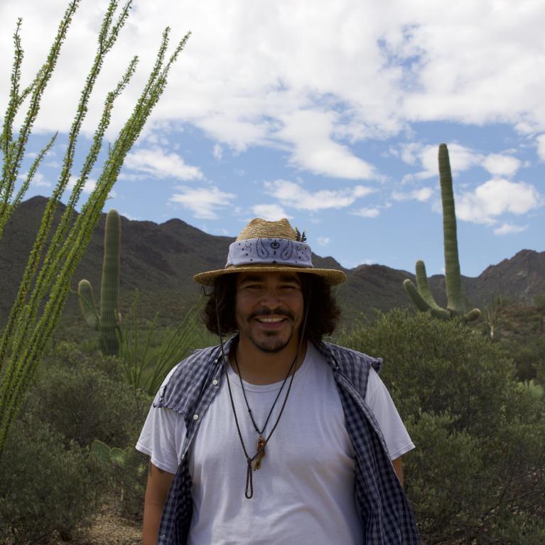 Isaac Silva wearing a hat and white shirt outside under a sunny sky with desert scenery in the background outside under a sunny sky with desert scenery in the background